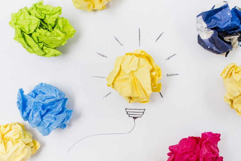 Zerknüllte farbige Papierkugeln sind auf einer weißen Fläche angeordnet. Eine gelbe Kugel befindet sich in der Mitte, umgeben von gezeichneten Linien, die an eine Glühbirne erinnern, während andere farbige Kugeln um sie herum verstreut sind.