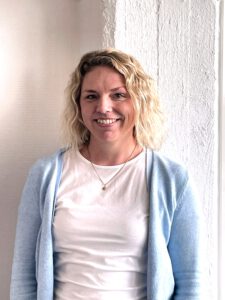 Eine Frau mit gewelltem blondem Haar, die eine hellblaue Strickjacke über einem weißen Hemd und eine Halskette mit Anhänger trägt, lächelt, während sie in einem Innenraum vor einer strukturierten weißen Wand steht.