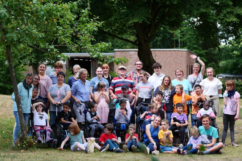 Eine bunt gemischte Gruppe von Kindern und Erwachsenen, von denen einige auf Rollstühle angewiesen sind, posiert und lächelt gemeinsam im Freien unter Bäumen, mit einem Backsteingebäude im Hintergrund.
